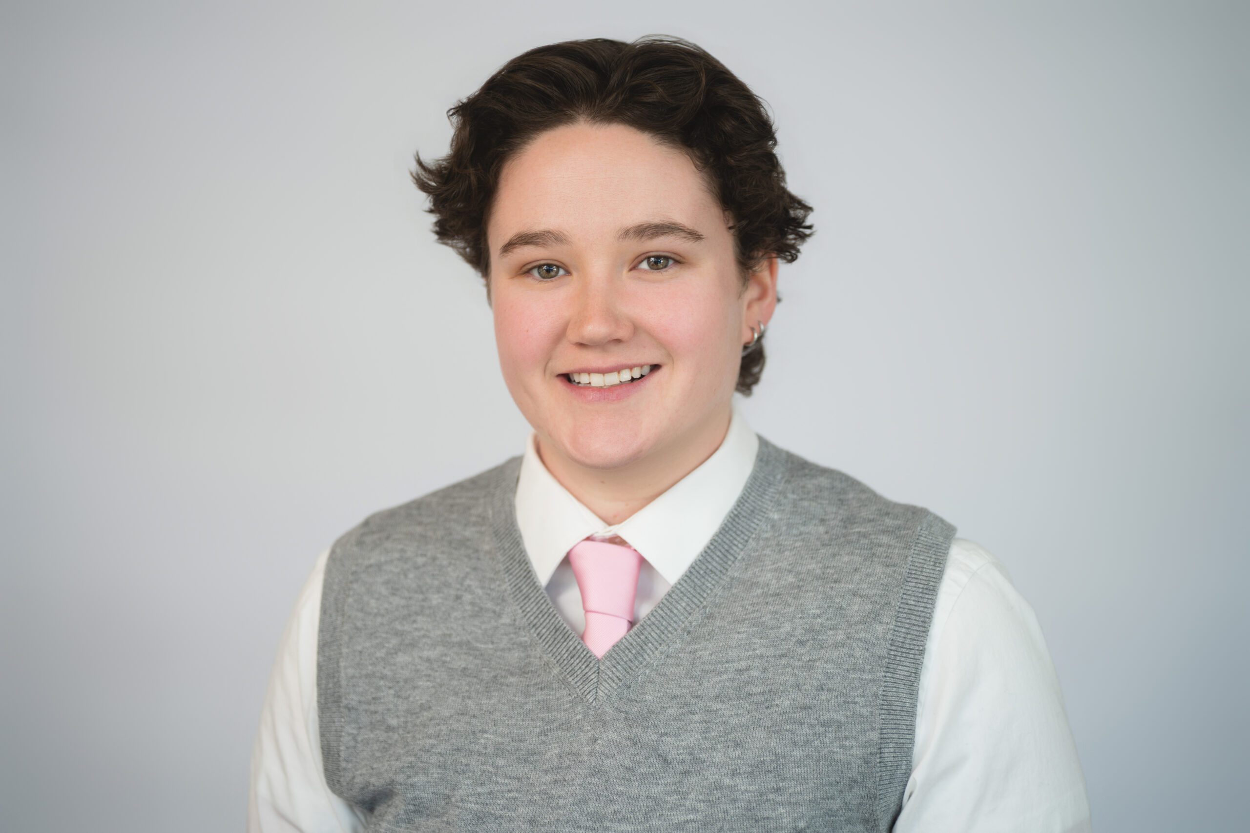 Jenna Headshot Person with short dark hair wearing a gray sweater vest, white shirt, and light pink tie, smiling in front of a plain light background.