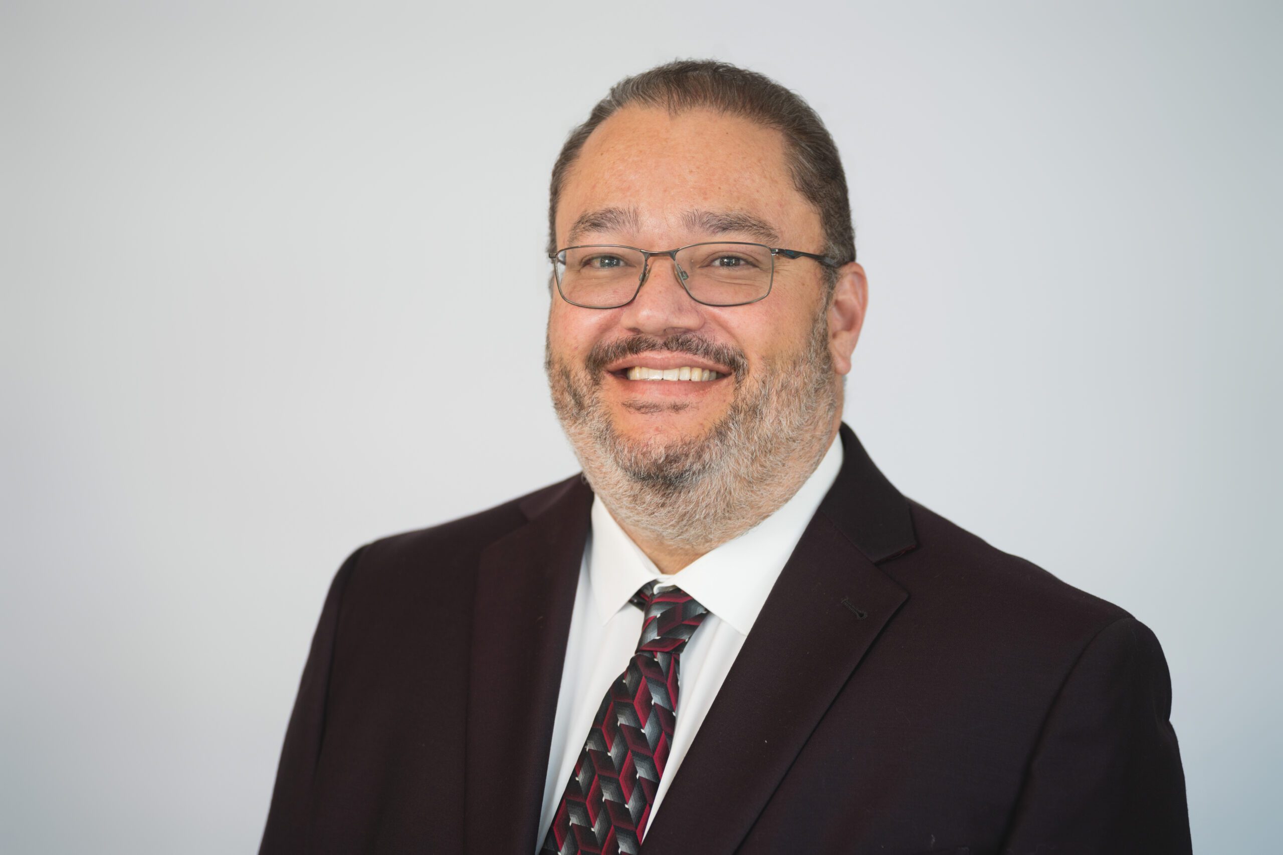 Ron Headshot Man wearing glasses, a dark suit, white shirt, and patterned tie, smiling in front of a plain light background.