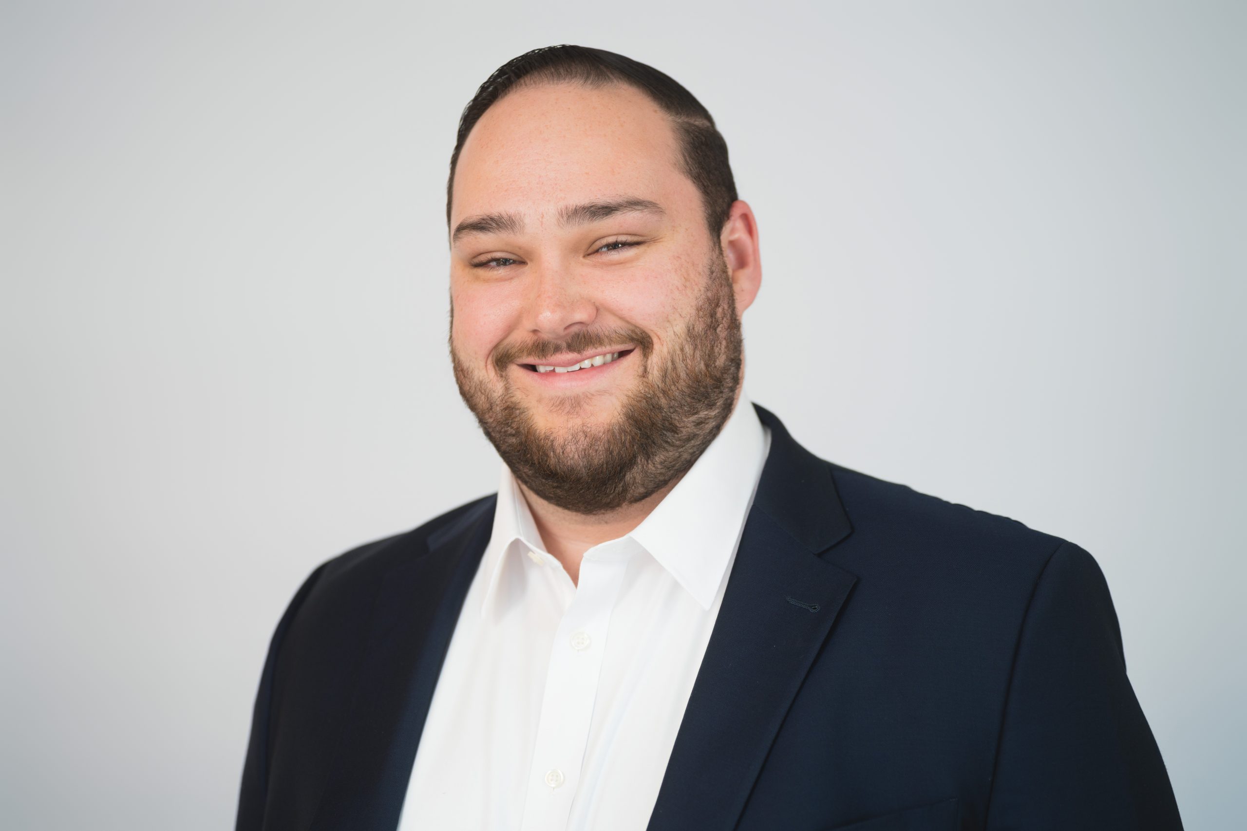 Ronnie Headshot 1 Man with short dark hair and a beard, wearing a white shirt and dark blazer, smiles in front of a plain light background.